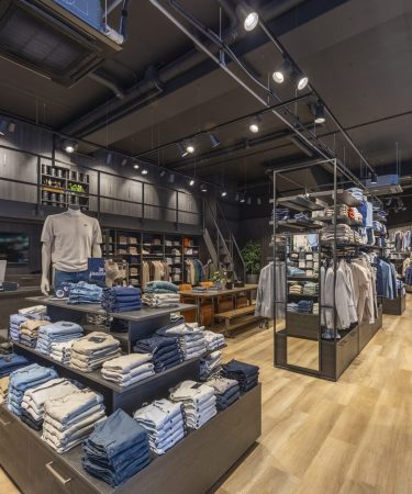 Brede winkelopstelling met nette kledingrekken gevuld met shirts, jassen en broeken in aardetinten.