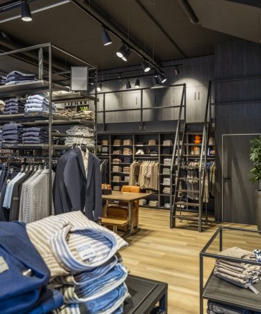 Winkelgedeelte met houten planken vol gevouwen kleding, een displaytafel, ladder en groene plant voor een warme uitstraling.