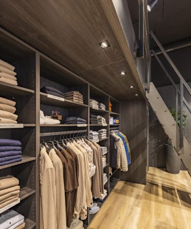 Lange winkelgang met donkere rekken vol gevouwen kleding en hangende jassen, sfeervol verlicht met spots.