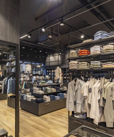 Brede blik op de winkel met meerdere rekken en planken vol shirts, truien en andere kledingstukken in een moderne setting.