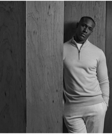 Zwart-wit portret van een man in een lichte trui met halve rits en lichte broek, leunend tegen een houten wand.