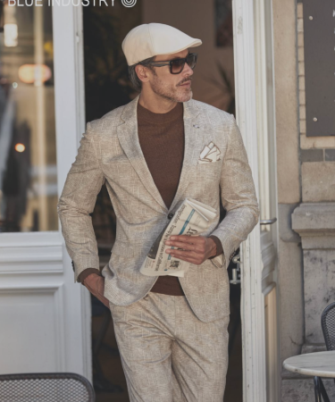 Man in een lichtbeige pak met coltrui en pet, staand in een deuropening en een krant vasthoudend, gefotografeerd in een stijlvolle stedelijke setting.