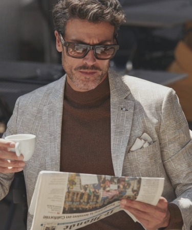 Man in een licht pak met coltrui en zonnebril, zittend op een terras terwijl hij een krant leest en een kopje koffie vasthoudt.