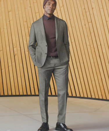 Model draagt een grijs herenpak met trui en overhemd, gefotografeerd voor een houten wand in een moderne setting.